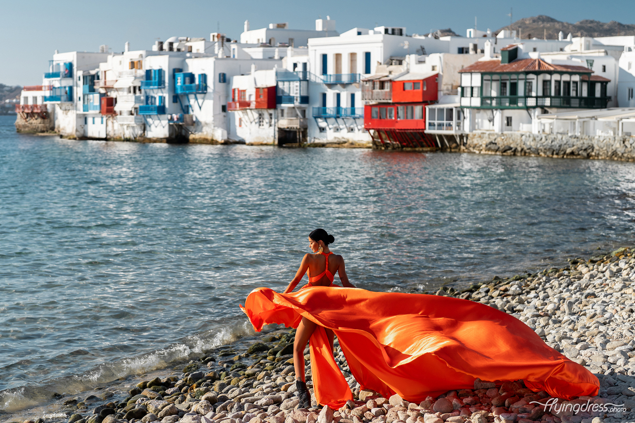 A woman in a vibrant orange dress gracefully stands on the rocky shore of Mykonos' Little Venice, with the colorful waterfront houses providing a picturesque backdrop.